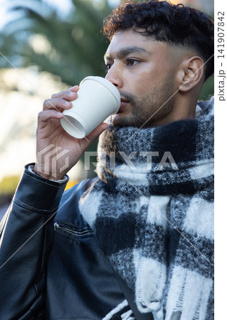 Man sipping from white disposable cup while wearing leather jacket and plaid scarf near palm trees Man sipping from white disposable cup while wearing leather jacket and plaid scarf near palm trees 141907842