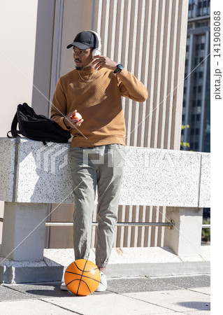 African American man leaning on terrace ledge, adjusting headset, holding apple, basketball by feet 141908088