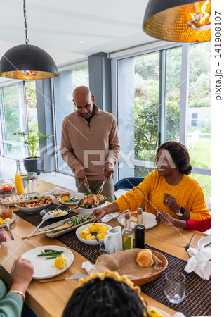 Family gathering around long wooden dining table at home, host carving roasted chicken 141908107