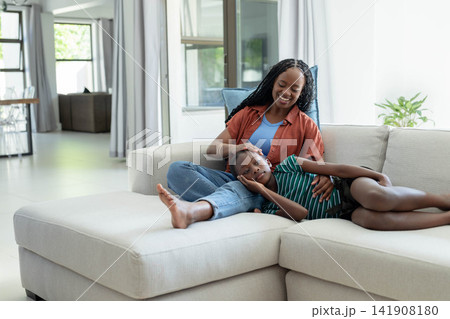 Mother and daughter lying on L-shaped sofa at home, woman in rust shirt caressing youth's head 141908180