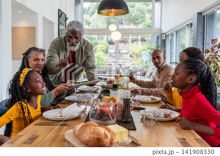 Multigenerational African American family gathering at wooden dining table at home, passing platter 141908330