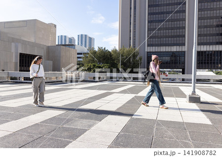 Two women crossing tiled urban plaza holding smartphone, cup and backpacks, casting long shadows 141908782