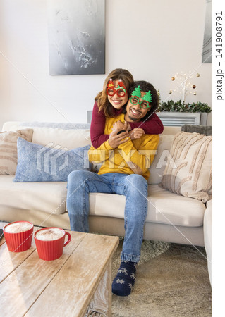 Couple hugging on beige sofa at home, wearing novelty glasses and holding smartphone near red mugs 141908791