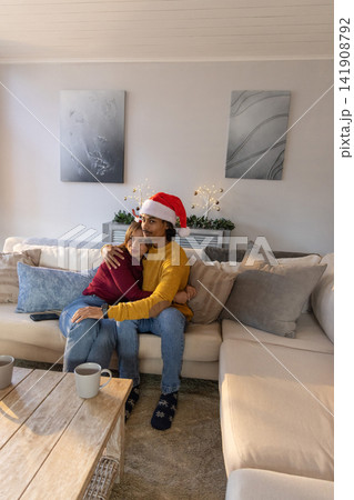 Couple cuddling on beige sectional sofa at home, wearing Santa hat beside coffee table with mugs Couple cuddling on beige sectional sofa at home, wearing Santa hat beside coffee table with mugs 141908792