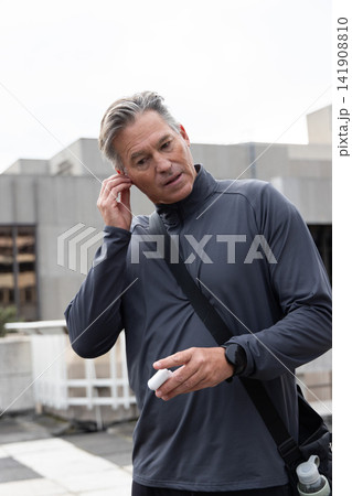 Senior man standing on rooftop wearing gray zip-up top inserting earbud and holding white case 141908810