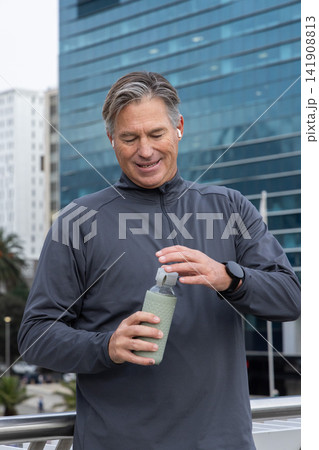 Middle-aged man standing on walkway railing in athletic jacket with earbuds opening reusable bottle Middle-aged man standing on walkway railing in athletic jacket with earbuds opening reusable bottle 141908813