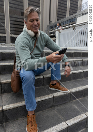 Senior man sitting on concrete steps checking smartphone while wearing headphones and crossbody bag 141908819