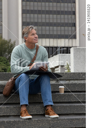 Middle-aged man sitting on steps city plaza, drawing in sketchbook, holding pencil, folio, bag, cup Middle-aged man sitting on steps city plaza, drawing in sketchbook, holding pencil, folio, bag, cup 141908820