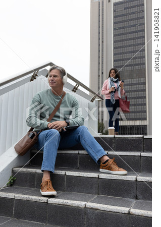 Commuters sitting on steps by office tower, man wearing headset using tablet, woman holding cup 141908821