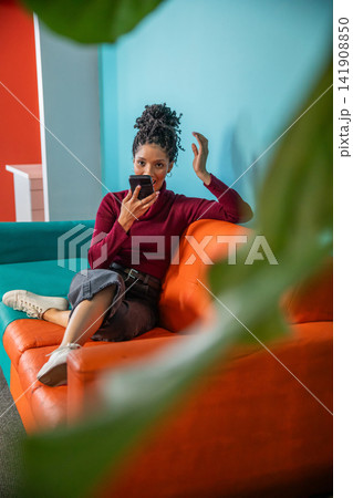 African American woman speaking into smartphone while sitting on orange sofa in colorful lounge African American woman speaking into smartphone while sitting on orange sofa in colorful lounge 141908850