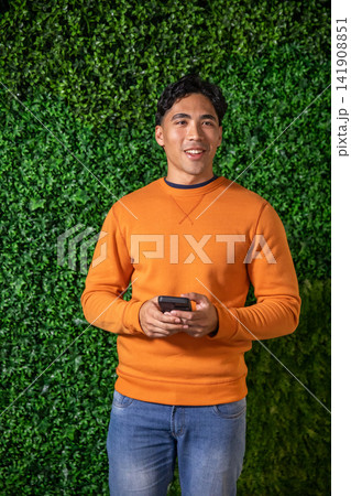 Adult Asian man standing in front of green hedge wall holding phone wearing orange sweatshirt jeans Adult Asian man standing in front of green hedge wall holding phone wearing orange sweatshirt jeans 141908851