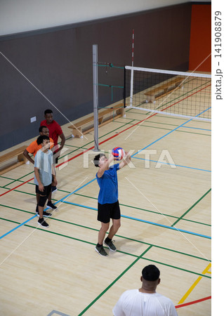 Diverse male team practicing at gym, teen in blue shirt sending red-white-blue volleyball overhead Diverse male team practicing at gym, teen in blue shirt sending red-white-blue volleyball overhead 141908879