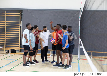 Diverse male volleyball team huddling near net post on gym court holding blue-white-red volleyball 141908885