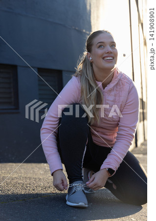 Female crouching, tying gray shoe on asphalt beside wall with pink jacket, smartwatch, copy space Female crouching, tying gray shoe on asphalt beside wall with pink jacket, smartwatch, copy space 141908901