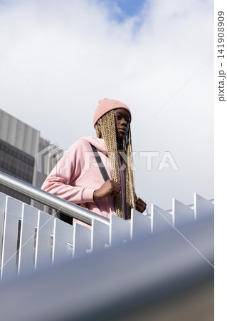 African American woman standing on stair wearing pink hoodie and beanie, holding backpack strap African American woman standing on stair wearing pink hoodie and beanie, holding backpack strap 141908909