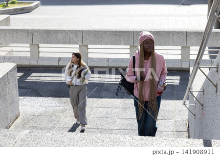 Diverse female students walking up campus stairs, carrying backpack, notebooks, coffee, copy space Diverse female students walking up campus stairs, carrying backpack, notebooks, coffee, copy space 141908911