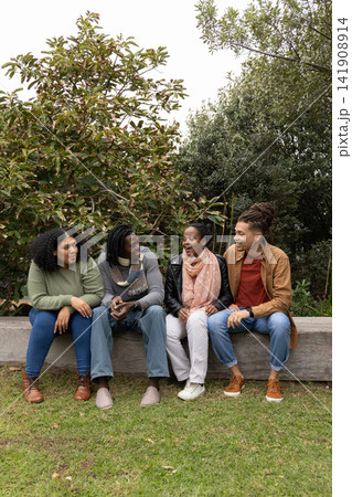 Diverse friends sitting on bench in park chatting, holding phone, wearing scarves and jeans 141908914