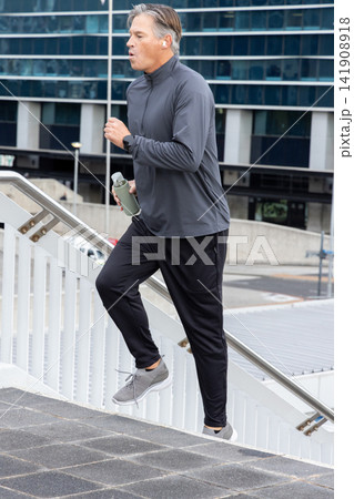 Middle-aged man climbing urban stairs in sportswear holding textured bottle, wearing earbuds, watch Middle-aged man climbing urban stairs in sportswear holding textured bottle, wearing earbuds, watch 141908918