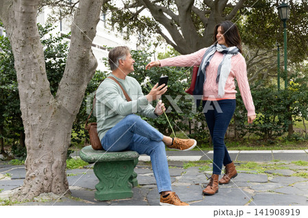 Couple exchanging daisy, man holding smartphone, woman reaching, scarf fluttering on park bench Couple exchanging daisy, man holding smartphone, woman reaching, scarf fluttering on park bench 141908919