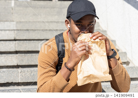 African American man sitting on concrete steps, eating from brown paper bag, wearing black backpack African American man sitting on concrete steps, eating from brown paper bag, wearing black backpack 141909491