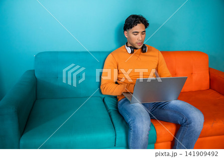 Man sitting on two-tone sofa wearing orange sweatshirt with headphones using laptop, copy space Man sitting on two-tone sofa wearing orange sweatshirt with headphones using laptop, copy space 141909492