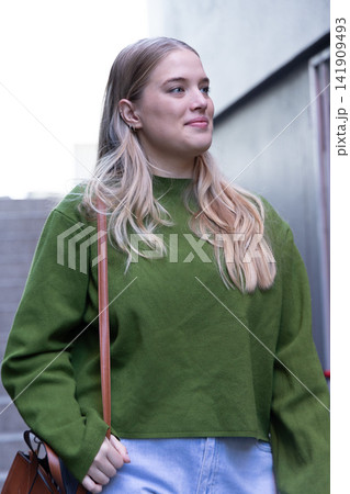 20s woman standing in stairwell by concrete wall, in green sweater and jeans, holding brown strap 141909493