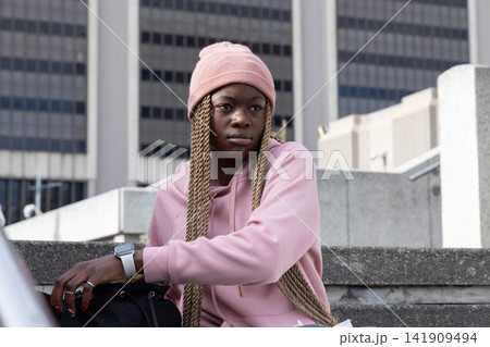 African American woman sitting on concrete steps in plaza in beanie leaning on bag with smartwatch 141909494