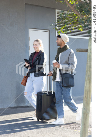 Couple walking beside grey facade, woman holding phone, man pulling black suitcase, sipping coffee 141909495
