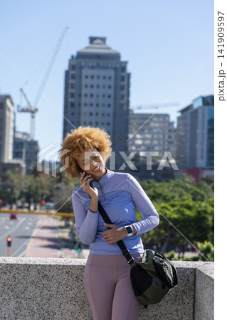 African American woman leaning on stone railing talking on phone, checking watch with bag in city African American woman leaning on stone railing talking on phone, checking watch with bag in city 141909597