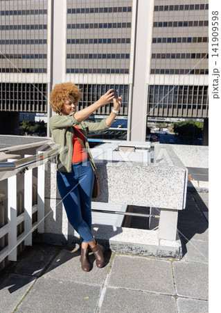African American woman leaning on parapet holding smartphone for selfie on rooftop near handrail African American woman leaning on parapet holding smartphone for selfie on rooftop near handrail 141909598