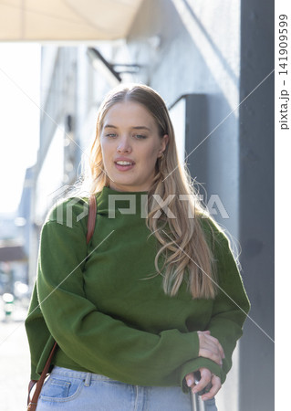 Woman walking on city sidewalk beside dark wall, wearing green sweater and holding metal handle Woman walking on city sidewalk beside dark wall, wearing green sweater and holding metal handle 141909599