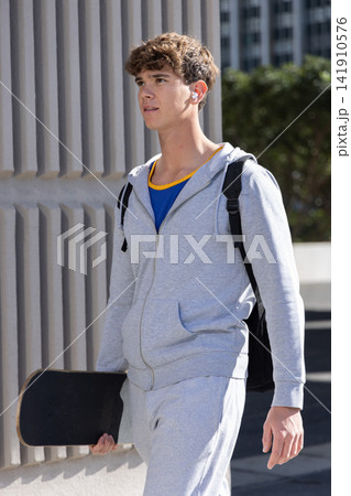 Adolescent male walking on sidewalk by ribbed wall carrying skateboard, wearing hoodie and backpack 141910576