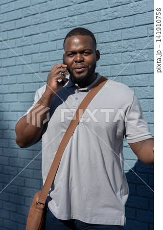 African American man 30s standing, talking on phone against blue brick wall with gray polo, satchel African American man 30s standing, talking on phone against blue brick wall with gray polo, satchel 141910758