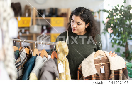 Stylish latina young woman standing in clothing shop and looking new coat or jacket made of high-quality material Stylish latina young woman standing in clothing shop and looking new coat or jacket made of high-quality material 141912108