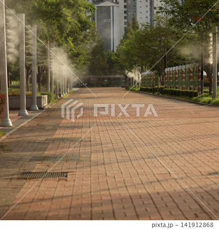Sunlit park walkway filled with cooling mist from outdoor water misters Sunlit park walkway filled with cooling mist from outdoor water misters 141912868
