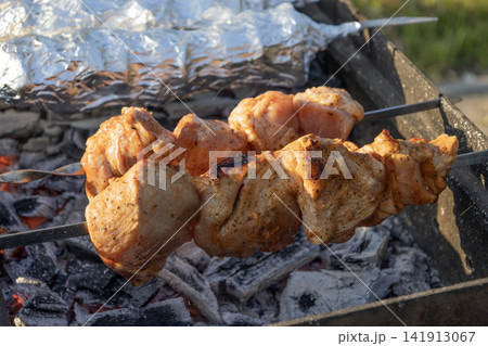 Delicious meat shashlik skewers and foil-wrapped food grilling on hot coals in a metal brazier outdoors. Summer picnic backyard barbecue cooking process with smoke and glowing charcoal embers. 141913067