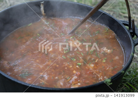 Delicious traditional Ukrainian red borscht soup cooking in a cast iron pot outdoors on fire, stirred with a ladle, showcasing fresh vegetables and rich broth for camping and nature travel food. Delicious traditional Ukrainian red borscht soup cooking in a cast iron pot outdoors on fire, stirred with a ladle, showcasing fresh vegetables and rich broth for camping and nature travel food. 141913099