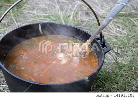 Delicious traditional Ukrainian red borscht soup cooking in a cast iron pot outdoors on fire, stirred with a ladle, showcasing fresh vegetables and rich broth for camping and nature travel food. Delicious traditional Ukrainian red borscht soup cooking in a cast iron pot outdoors on fire, stirred with a ladle, showcasing fresh vegetables and rich broth for camping and nature travel food. 141913103