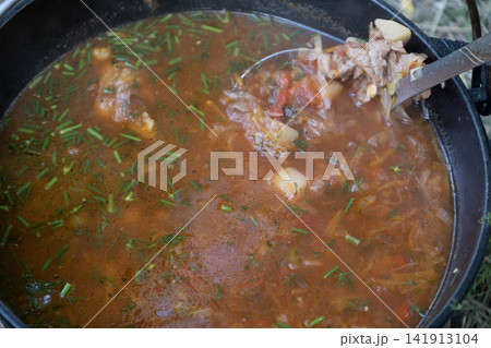 Delicious traditional Ukrainian red borscht soup cooking in a cast iron pot outdoors on fire, stirred with a ladle, showcasing fresh vegetables and rich broth for camping and nature travel food. Delicious traditional Ukrainian red borscht soup cooking in a cast iron pot outdoors on fire, stirred with a ladle, showcasing fresh vegetables and rich broth for camping and nature travel food. 141913104