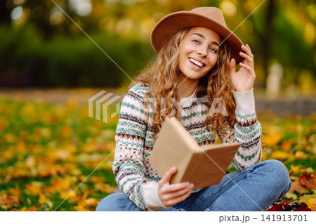 Young woman in hat sits on a plaid in autumn park and reads a book. Concept of nature, education. Young woman in hat sits on a plaid in autumn park and reads a book. Concept of nature, education. 141913701
