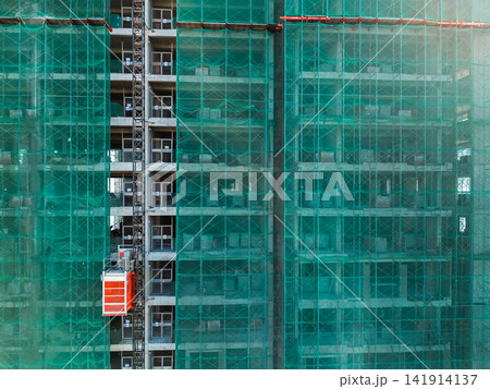 High-rise building under construction with green safety mesh and external lift elevator, Modern urban development scene showing scaffolding structure and unfinished concrete facade High-rise building under construction with green safety mesh and external lift elevator, Modern urban development scene showing scaffolding structure and unfinished concrete facade 141914137