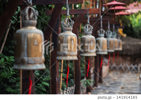 Hanging temple bells with red ropes along wooden walkway in Thailand, Traditional Buddhist metal bells in row with perspective and spiritual atmosphere 141914165