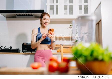 Fitness woman reading recipe while preparing healthy smoothie with fresh vegetables in modern kitchen. Healthy lifestyle, nutrition, home cooking 141914537
