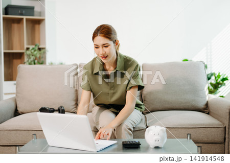 Smart financial management. Young Asian woman planning budget and savings using laptop, calculator, and piggy bank in a bright living room 141914548