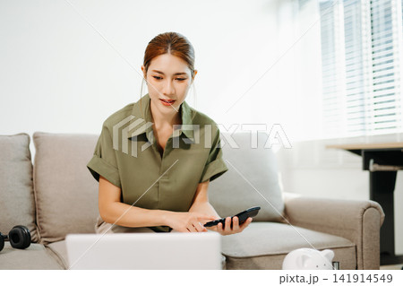 Smart financial management. Young Asian woman planning budget and savings using laptop, calculator, and piggy bank in a bright living room Smart financial management. Young Asian woman planning budget and savings using laptop, calculator, and piggy bank in a bright living room 141914549