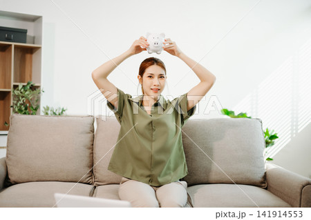 Smart financial management. Young Asian woman planning budget and savings using laptop, calculator, and piggy bank in a bright living room Smart financial management. Young Asian woman planning budget and savings using laptop, calculator, and piggy bank in a bright living room 141914553