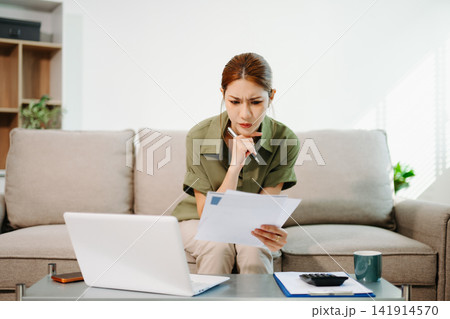Financial stress and debt management. Worried Asian woman analyzing domestic bills and expenses with laptop and calculator in her home living room. Financial stress and debt management. Worried Asian woman analyzing domestic bills and expenses with laptop and calculator in her home living room. 141914570