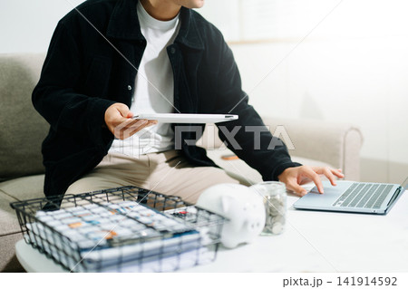 Close up of a white piggy bank and coin jar on a table while a person manages personal finance and budgeting in the background. Close up of a white piggy bank and coin jar on a table while a person manages personal finance and budgeting in the background. 141914592