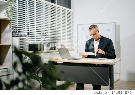 Experienced senior male executive reviewing reports and using a calculator at his office desk Experienced senior male executive reviewing reports and using a calculator at his office desk 141914879