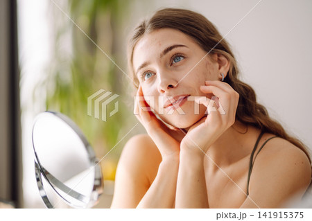 Young woman with problem skin looking into mirror. Inflammation on face, problem skin. Beauty care. Young woman with problem skin looking into mirror. Inflammation on face, problem skin. Beauty care. 141915375
